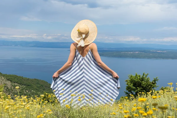 Visão traseira da jovem mulher vestindo vestido de verão listrado e chapéu de palha em pé em super flor de flores silvestres, relaxante enquanto desfruta de bela vista da natureza do mar Adriático, Croácia — Fotografia de Stock