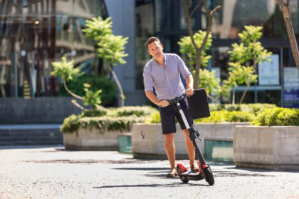 Jonge casual actieve sportieve zakenman in het centrum van de stad, het dragen van shirt en shorts, het vasthouden van laptop tas, rijden naar het werk op elektrische scooter op een hete zomerdag — Stockfoto
