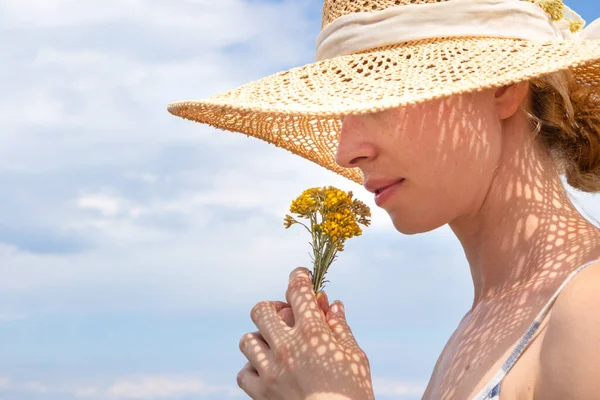 Porträtt av ung vacker glad kvinna bär halm sol hatt, luktar liten bukett gula vilda blommor, mot blå sommar himmel — Stockfoto