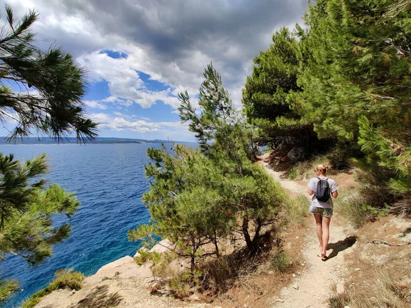 Fiatal aktív feamle turista visel kis hátizsák sétál a tengerparti ösvényen fenyőfák között keresi a távoli öböl úszni egyedül békében a tengerparton Horvátországban. Utazási és kalandkoncepció — Stock Fotó