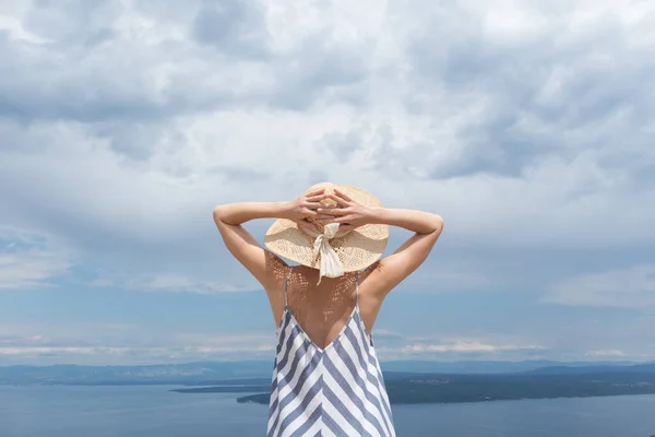 Bakifrån av ung kvinna bär randig sommar klänning och halm hatt stående i super blomning av vilda blommor, avkopplande samtidigt som den vackra utsikten över Adriatiska havet natur, Kroatien — Stockfoto