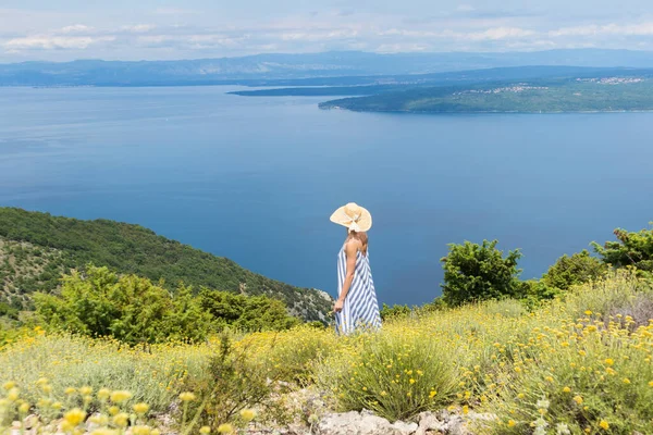 Vista trasera de la mujer joven con vestido de verano a rayas y sombrero de paja de pie en super floración de flores silvestres, relajante mientras disfruta de una hermosa vista de la naturaleza del mar Adriático, Croacia — Foto de Stock