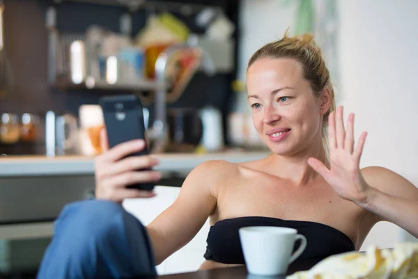 Unga leende glad nöjd kvinna inomhus i köket med hjälp av sociala medier på mobiltelefon för att chatta och hålla kontakten med sina nära och kära. Stanna hemma, social distanserande livsstil. — Stockfoto