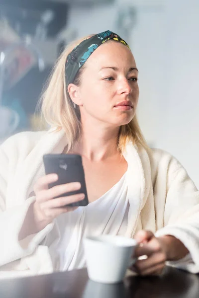 Hermosa mujer caucásica en casa, sintiéndose cómoda usando albornoz blanco, tomando algún tiempo para sí misma, bebiendo café de la mañana y leyendo noticias en el dispositivo de teléfono móvil por la mañana — Foto de Stock