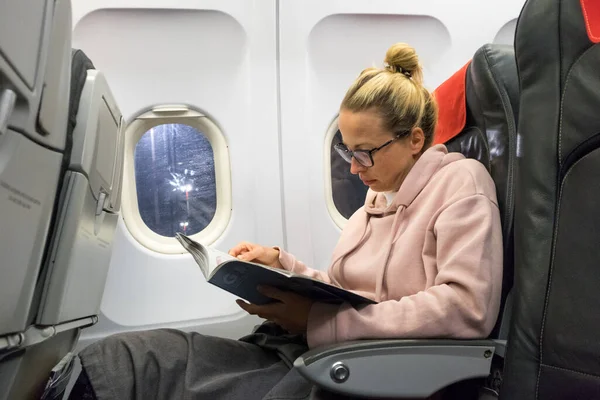 Jovem mulher casual ler revista e ouvir música no avião. Viajante fêmea lendo sentado na cabine de passageiros. — Fotografia de Stock