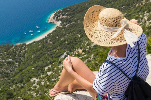 여름휴가 때 활동적 인 운동을 하는 한 여성이 루 벤체 마을의 오래 된 석조 벽에 앉아, 밀짚 모자를 쓰고 크로아티아의크 레아 섬의 아름다운 해안 풍경을 즐기고 있는 해변 배낭을 메고 있다 — 스톡 사진