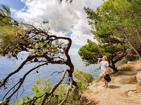 Joven turista activo tomando un descanso, bebiendo agua, usando una pequeña mochila mientras camina por el sendero costero entre pinos en busca de una cala remota para nadar solo en paz en la costa en Croacia — Foto de Stock