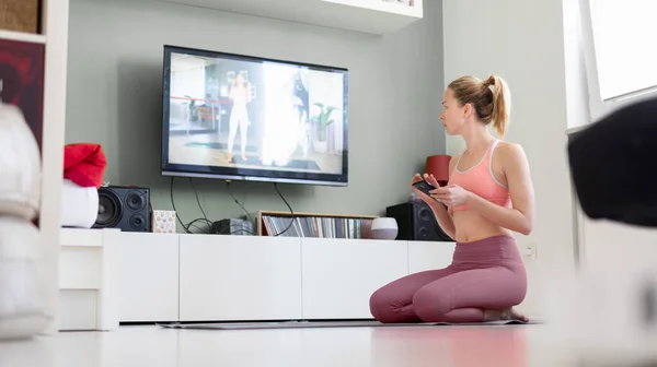 Atrakcyjna sportowa kobieta ćwicząca w domu, ćwicząca pilates przed telewizorem w swoim małym mieszkaniu. Oddalenie się od społeczeństwa. Bądź zdrowy i pozostań w domu podczas pandemii wirusa korony — Zdjęcie stockowe