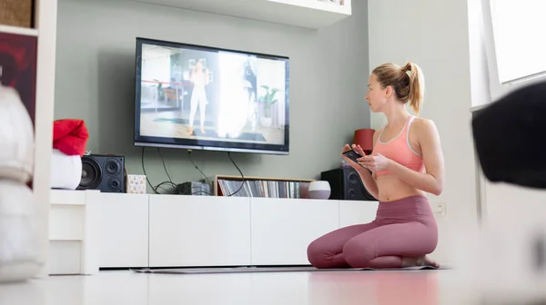 Atractiva mujer deportiva haciendo ejercicio en casa, haciendo ejercicios de pilates frente a la televisión en su pequeño apartamento de estudio. Distanciamiento social. Manténgase saludable y permanezca en casa durante la pandemia del virus corona — Foto de Stock