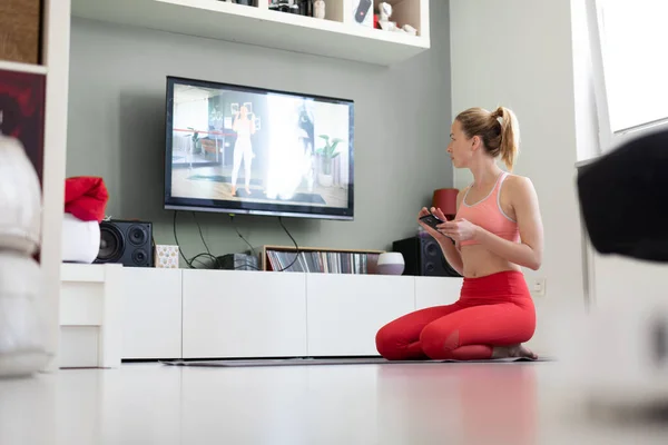 Atractiva mujer deportiva haciendo ejercicio en casa, haciendo ejercicios de pilates frente a la televisión en su pequeño apartamento de estudio. Distanciamiento social. Manténgase saludable y permanezca en casa durante la pandemia del virus corona — Foto de Stock