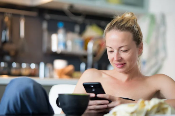 Jeune femme heureuse souriante à l'intérieur de la cuisine à la maison en utilisant les médias sociaux sur le téléphone mobile pour discuter et rester connecté avec ses proches. Rester à la maison, mode de vie social distanciant. — Photo