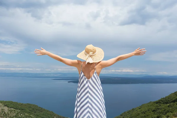 Vista trasera de la mujer joven con vestido de verano a rayas y sombrero de paja de pie en super floración de flores silvestres, relajarse con las manos hacia el cielo, disfrutando de una hermosa vista de la naturaleza del mar Adriático, Croacia —  Fotos de Stock