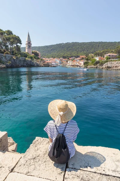 Mulher viajante vestindo chapéu de verão de palha e mochila, sentado na borda do cais de pedra, desfrutando de bela vista panorâmica de Veli Losinj, ilha de Losinj, Croácia. — Fotografia de Stock