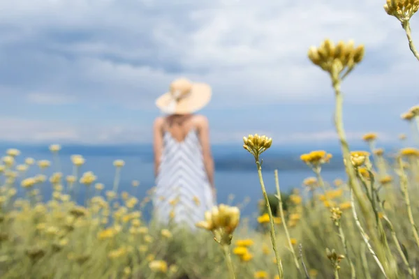 Vista trasera de la mujer joven con vestido de verano a rayas y sombrero de paja de pie en super floración de flores silvestres, relajante mientras disfruta de una hermosa vista de la naturaleza del mar Adriático, Croacia —  Fotos de Stock