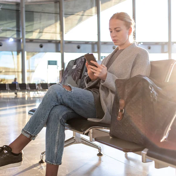 Mujer joven rubia casual usando su teléfono celular mientras espera para abordar un avión en las puertas de salida del aeropuerto. — Foto de Stock