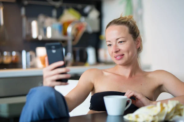 Unga leende glad nöjd kvinna inomhus i köket med hjälp av sociala medier på mobiltelefon för att chatta och hålla kontakten med sina nära och kära. Stanna hemma, social distanserande livsstil. — Stockfoto