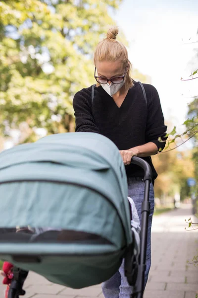 忧心忡忡的年轻妈妈带着婴儿车走在空荡荡的街道上，戴着医疗面罩，以保护她免受科罗纳病毒的侵害。日冕病毒大流行期间的社会疏离生活 — 图库照片