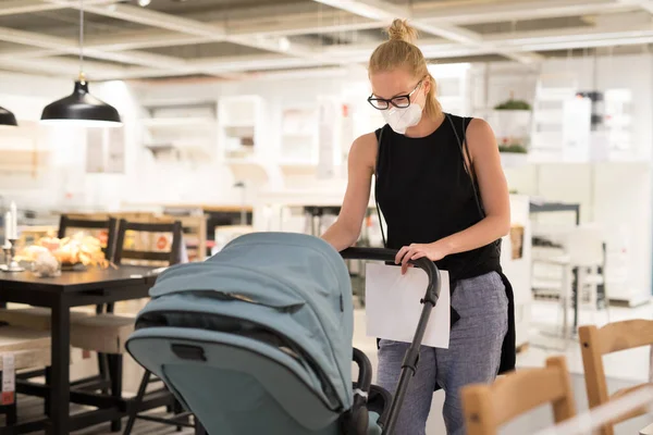 Young mom with newborn in stroller shopping at retail furniture and home accessories store wearing protective medical face mask to prevent spreading of corona virus. New normal during covid epidemic