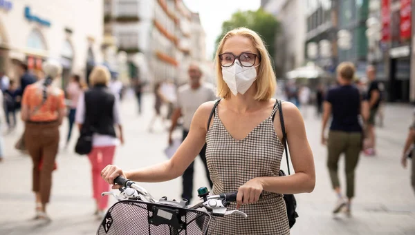 Woman walking by her bicycle on pedestrian city street wearing medical face mask in public to prevent spreading of corona virus. New normal during covid epidemic.