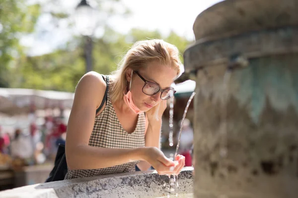 Törstig ung casual cucasian kvinna bär medicinsk ansiktsmask dricksvatten från offentliga staden fontän på en varm sommardag. Nya sociala normer under covid epidemi — Stockfoto