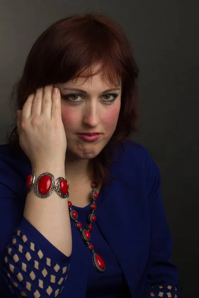 Portrait of a full grown woman, brunette, in the Studio on a gray background. In a blue dress and red jewelry