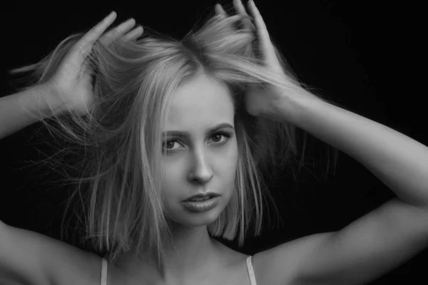 black-and-white portrait of a blonde on a black background. in a white tank top with straps. hand-ruffled hair