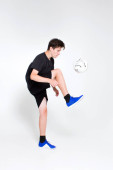 Mladý chlapec v černém sportovním tričku a šortkách, modrých teniskách, vlacích a hraje si s fotbalovým míčem.