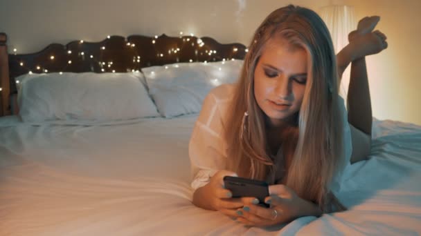 Mooi blond meisje gebruik haar mobiele telefoon terwijl liggend op bed. Internet dating — Stockvideo