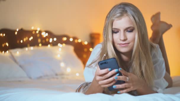 Mooi blond meisje gebruik haar mobiele telefoon terwijl liggend op bed. Internet dating — Stockvideo