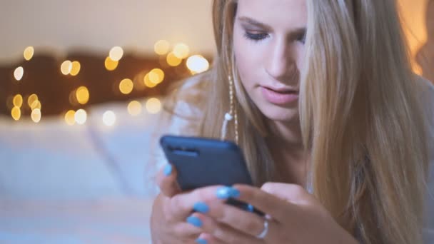 Schöne blonde Mädchen benutzen ihr Handy, während sie auf dem Bett liegen. Internet-Dating aus nächster Nähe — Stockvideo