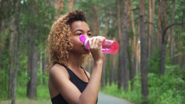 Svart kvinna dricka från Rosa flaska. Porträtt av svart ung kvinna tar paus efter jogging — Stockvideo