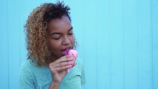 Jovem menina negra comendo sorvete rosa e sorrindo no fundo da parede azul — Vídeo de Stock