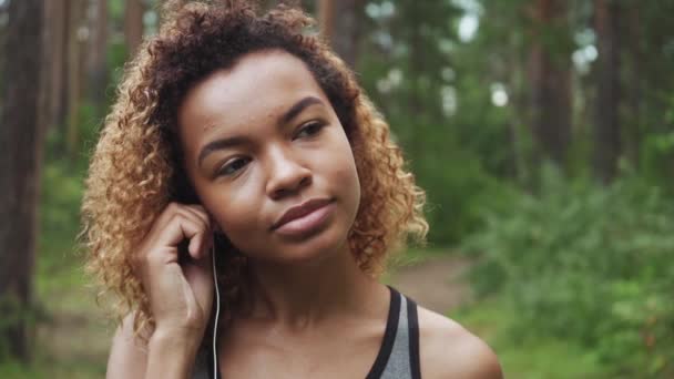 Mladá krásná afroameričanka s kudrnatými vlasy klade na sluchátka před běhání v lese — Stock video