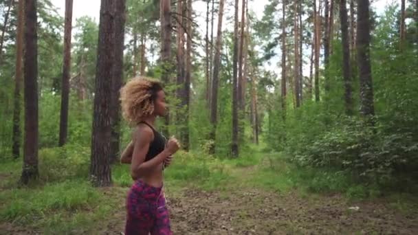 Молодая красивая афроамериканка с вьющимися волосами, бегущая с музыкой в лесу — стоковое видео