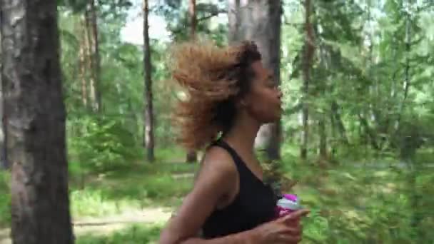 Jovem bela mulher afro-americana com cabelo encaracolado correndo com garrafa rosa em uma floresta — Vídeo de Stock