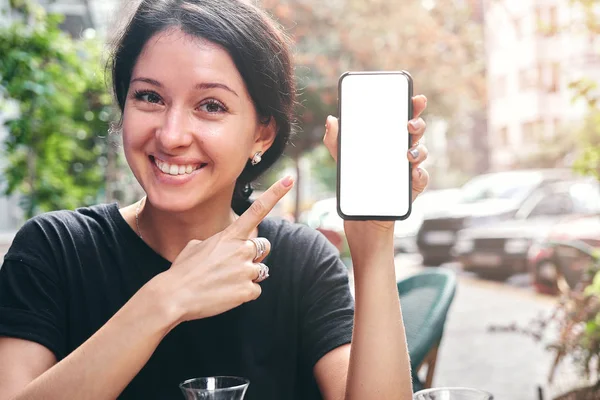 Mulher bonita feliz segurando e mostrando telefone celular com tela branca em branco no café — Fotografia de Stock