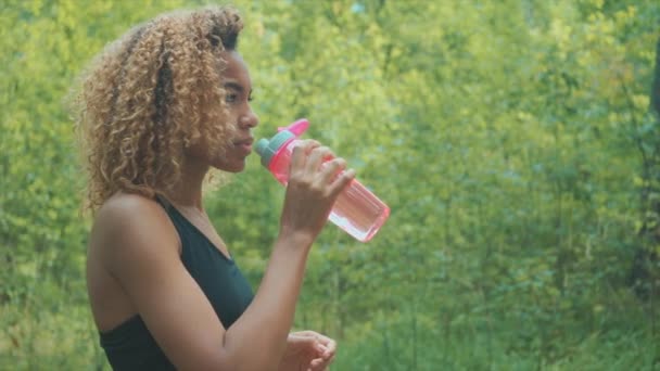 Mujer negra bebiendo de una botella rosa. Retrato de una joven negra tomando un descanso mientras trota — Vídeos de Stock