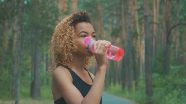 Zwarte vrouw uit roze fles drinken. Portret van zwarte jonge vrouw nemen van onderbreking tijdens het joggen — Stockvideo