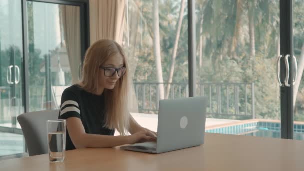 Блондинка печатает на своем ноутбуке за столом с бассейном за окном. внештатная концепция — стоковое видео