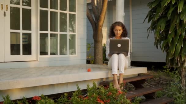 Eine junge, schöne Freiberuflerin sitzt auf der Terrasse mit einem Laptop im Hintergrund ihres Hauses — Stockvideo