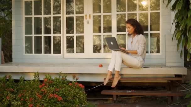 Μια νεαρή όμορφη γυναίκα ελεύθερος επαγγελματίας κάθεται στη βεράντα με ένα φορητό υπολογιστή στο φόντο του σπιτιού της — Αρχείο Βίντεο