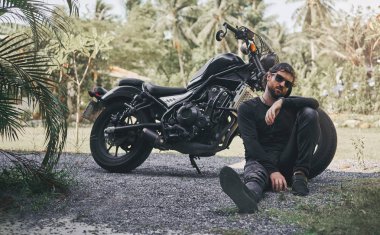 Siyah giyim yakışıklı bisikletçi adam klasik tarzı kafe yarışçı motosiklet yakınında oturup. özel yapım motosiklet