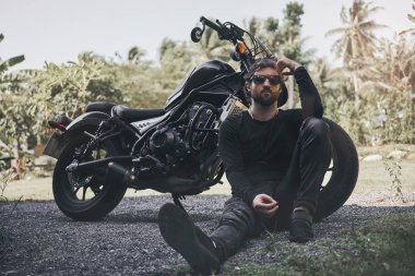 Siyah giyim yakışıklı bisikletçi adam klasik tarzı kafe yarışçı motosiklet yakınında oturup. özel yapım motosiklet