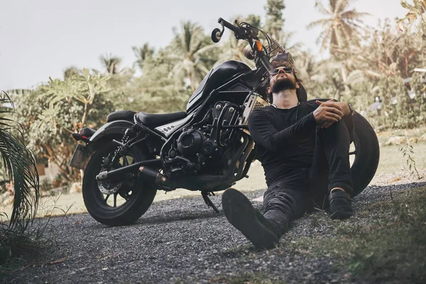 Siyah giyim yakışıklı bisikletçi adam klasik tarzı kafe yarışçı motosiklet yakınında oturup. özel yapım motosiklet