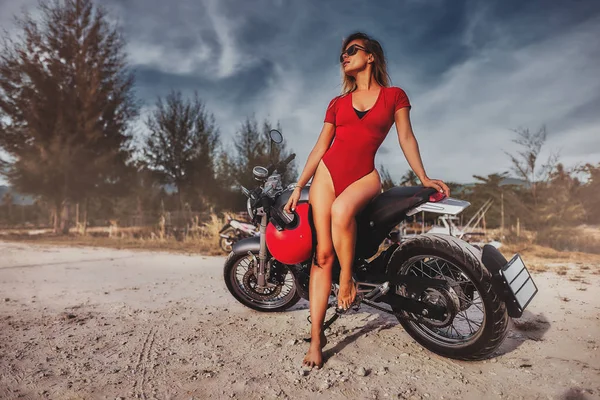 Outdoor lifestyle portrait of young sexy woman in red swimsuit sitting on a vintage custom motorcycle