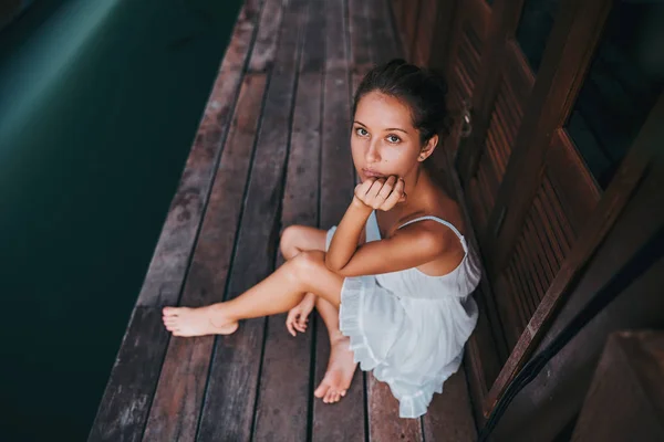 Piękna dziewczyna w białej sukni siedzi na drewnianej podłodze w pobliżu błękitnego jeziora i patrzy na kamerę — Zdjęcie stockowe