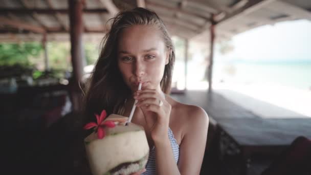 Žena popíná z kokosového ořechu pod vilou na pláži a dívá se přímo na kameru — Stock video