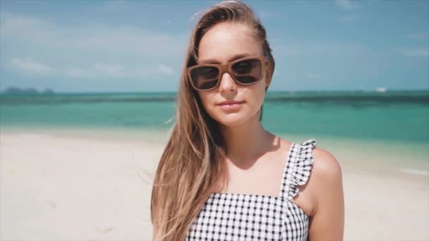 Mujer con gafas de sol de pie en la playa de arena mirando directamente a la cámara — Vídeo de stock