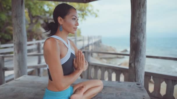 Mujer sentada bajo villa y meditando con las piernas cruzadas cerca del océano — Vídeos de Stock