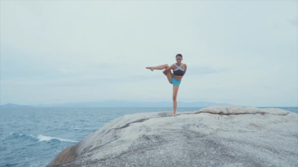 Mujer de pie sobre una pierna y meditando sobre rocas cerca del océano — Vídeos de Stock